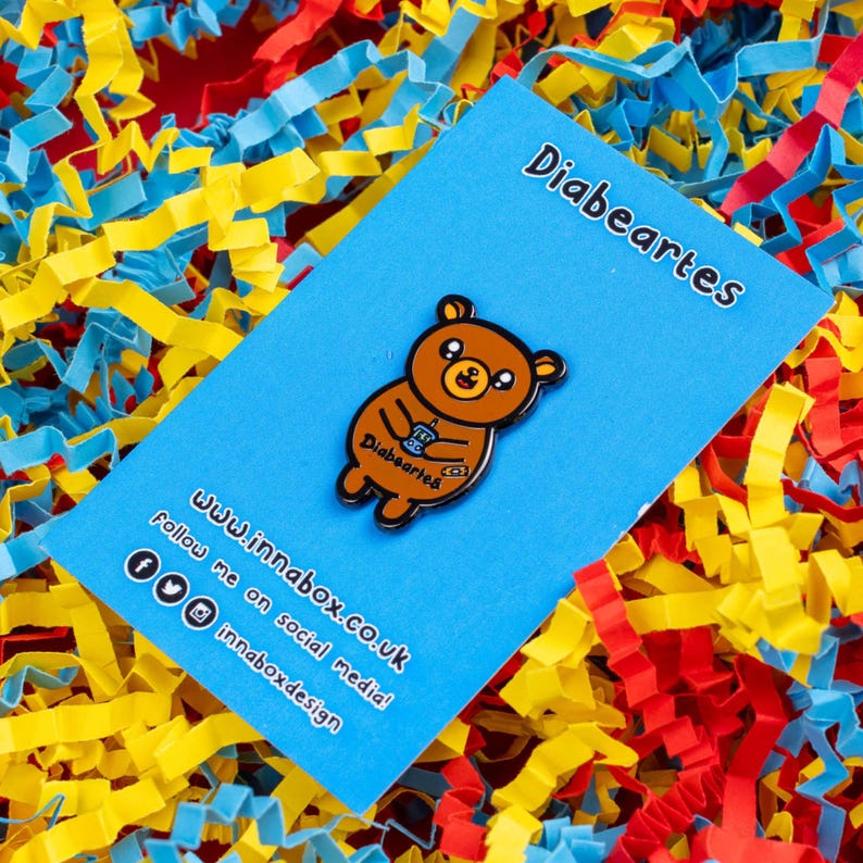 Diabeartes Enamel Pin - Diabetes on blue backing card on yellow, red & blue card. Smiley brown bear with a blood glucose reader & a plaster on its arm, across its tummy reads diabeartes in black. Raising awareness for type 1 & type 2 diabetes.
