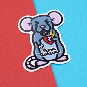 May include: A grey cartoon rat with a pink nose and ears is holding a red heart with a yellow lightning bolt through it. The rat is looking up with a surprised expression. The text "Panic Rattack" is written below the rat.