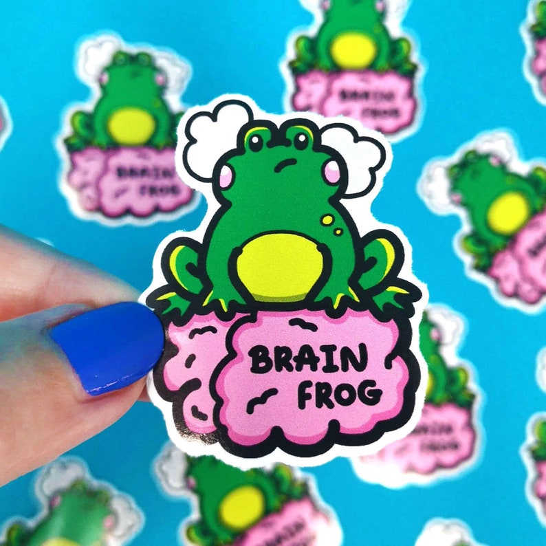Brain Frog Sticker - Brain Fog being held above multiple frog stickers over a blue background. A confused frog with pink cheeks & 2 grey clouds above. Sat on a pink brain with text reading Brain Frog. Created to raise awareness for brain fog.