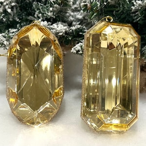 Op de afbeelding: Twee goudkleurige kerstversieringen met facetten. Eén is ovaal, de andere rechthoekig. Beide hebben een glitterende gouden rand en een lus om op te hangen. Ze staan tegen een achtergrond van een besneeuwde kerstboom.