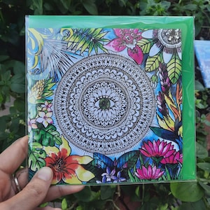 May include: A square greeting card with a black and white mandala design and an eye motif, framed by vibrant floral and foliage illustrations. The card is encased in a green cellophane wrapper.