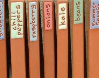 Op de afbeelding: Een verzameling terracotta gekleurde tuinmarkeringen met witte labels. Elk label toont de namen van verschillende groenten en fruit, waaronder "chili peppers" en "lettuce". De markeringen zijn in een rij gerangschikt, ideaal voor het organiseren van een tuin.