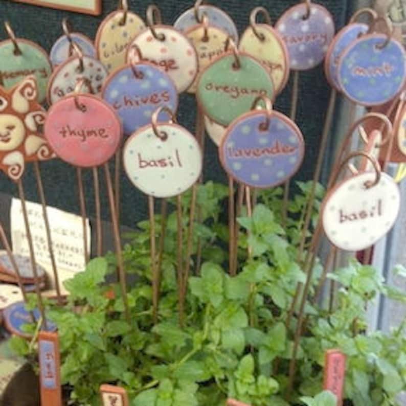 May include: A collection of ceramic garden markers with different colored backgrounds and handwritten names of herbs, including basil, thyme, chives, oregano, and lavender.