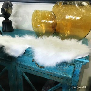 May include: White faux fur runner on a teal blue dresser with two gold glass vases. The runner is soft and fluffy, and the dresser has a distressed finish. The text "Fur Accents" is visible in the bottom right corner of the image.