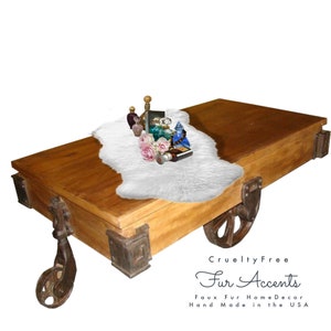 May include: A wooden coffee table with wheels, featuring a white faux fur rug and a collection of decorative bottles and boxes. The table has a rustic, industrial design with metal accents. The text "Cruelty Free Fur Accents" is visible below the table.  Faux Fur Home Decor Hand Made in the USA.