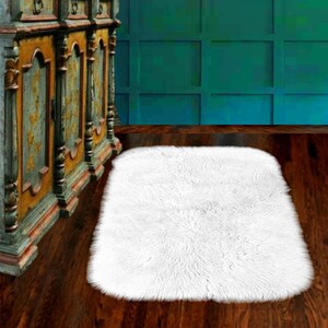 May include: A white fluffy rectangular rug with a slightly rounded edge. The rug is on a dark wood floor in front of a teal green wall.