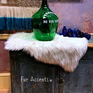 Puede incluir: Una alfombra de piel sintética blanca se encuentra sobre un gabinete de madera oscura con las palabras "Fur Accents" escritas en la parte delantera. Una gran botella de vidrio verde con las palabras "De Vin Vieux" escritas en ella se encuentra sobre la alfombra. Varias pequeñas botellas de vidrio azul están esparcidas alrededor de la botella grande.