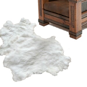 Op de afbeelding: Witte nep bontkleed met een zachte, pluche textuur. Het kleed ligt op een witte achtergrond met een houten stoel op de achtergrond.