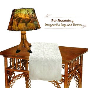 May include: A white faux fur table runner on a wooden table with a lamp featuring a deer print lampshade. The lamp has a wooden base with antlers. The text "Fur Accents® Designer Fur Rugs and Throws" is visible in the background.