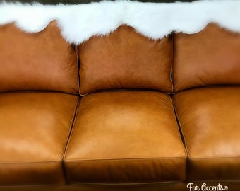 Könnte beinhalten: Ein braunes Ledersofa mit drei Kissen und einem weißen Kunstfellüberwurf, der über die Rückenlehne gelegt ist. Das Sofa hat eine gesteppte Rückenlehne und einen leicht abgenutzten Look. Der Text "Fur Accents®" ist in der unteren rechten Ecke des Bildes sichtbar.