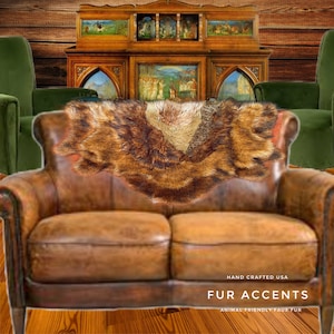 May include: A brown and tan faux fur throw blanket draped over a brown leather sofa. The throw has a textured pattern and is labeled "Fur Accents" and "Animal Friendly Faux Fur". The background includes green armchairs and a wooden cabinet.