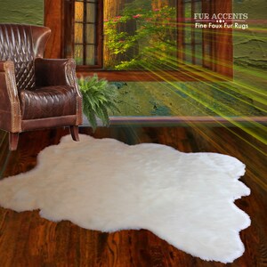 Op de afbeelding: Witte nep-bont vloerkleed op een houten vloer. Het kleed is gevormd als een schapenvacht en heeft een zachte, pluche textuur. Het kleed ligt voor een leren fauteuil en een potplant.
