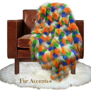 Op de afbeelding: Een kleurrijk nep bont deken met een patchwork ontwerp in tinten van oranje, groen, blauw, wit en roze. Het deken is gedrapeerd over een bruine leren fauteuil en een wit nep bont kleed. De tekst "Fur Accents®" is zichtbaar op het kleed.