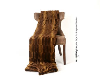 May include: A brown faux fur throw blanket draped over a brown upholstered chair. The blanket has a rectangular pattern with varying shades of brown. The chair has wooden legs and decorative nailhead trim.