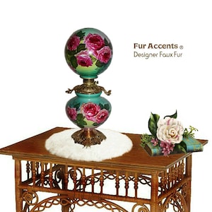May include: A vintage green glass lamp with a rose pattern on the shade and base. The lamp is sitting on a white fur table runner on a wooden table with a woven base.