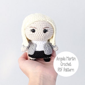 May include: Crocheted amigurumi doll of a woman with blonde hair, wearing a gray jacket, white shirt, and black skirt. The doll is holding a small white circle with the text "Angela Martin Crochet PDF Pattern".