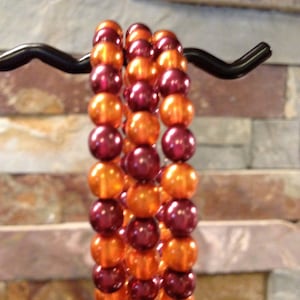 May include: Three beaded bracelets with alternating rows of orange and burgundy beads. The bracelets are displayed on a black metal hook against a textured brick wall background. The beads are round and have a glossy finish.
