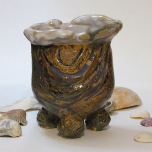 May include: A brown and green ceramic pot with a white glaze and three legs. The pot has a unique, organic shape and is decorated with a swirling pattern.