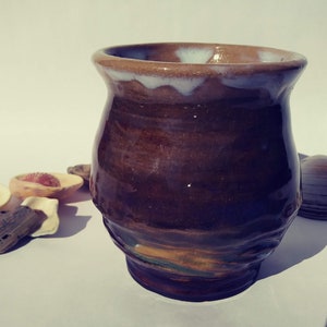 May include: A brown ceramic mug with a white rim and a glaze that creates a layered effect. The mug has a rounded body and a slightly flared lip.