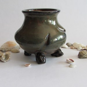 May include: A small, green ceramic pot with three black feet. The pot is sitting on a white surface with seashells scattered around it.