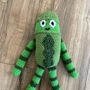 May include: A green crocheted plush toy with a striped pattern. The toy has a green hat and a smiling face with white eyes.