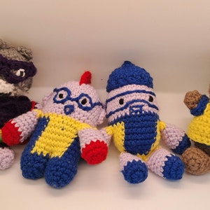 May include: Four crocheted stuffed toys, including a gray wolf wearing a purple mask, a purple and yellow robot with red hands, a blue and yellow robot with a blue hat, and a brown bear wearing a yellow shirt with a red star.