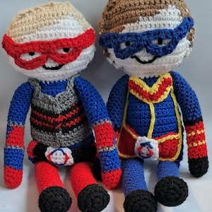 May include: Two handmade crochet dolls, each with a superhero theme. One doll has a tan head with a red mask, a gray and black vest, and red and blue accents. The other doll has a brown head with a blue mask and a blue, red, and yellow outfit.