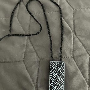 May include: A black necklace with a rectangular pendant. The pendant is white with a black woven pattern. The necklace cord is also black and appears to be made of a woven material. The necklace is displayed on a gray surface.