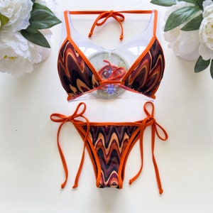 May include: Orange and brown bikini with a wavy pattern. The top is a triangle style with adjustable straps and ties at the neck. The bottom is a cheeky style with ties on the sides.