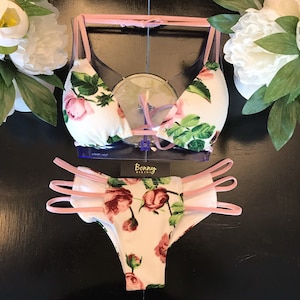 Peut inclure: Un bikini à imprimé floral blanc et rose avec un haut halter et un bas taille haute. Le bikini est doté d'une bordure rose et est fabriqué dans un tissu doux et extensible. Le bikini est présenté sur un mannequin.