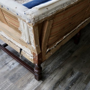 May include: Close-up of the underside of a vintage sofa with exposed wooden frame and fabric. The sofa has a dark blue fabric covering and a brown and white striped fabric lining. The sofa has four wooden legs with turned details.