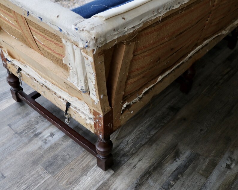 May include: Close-up of the underside of a vintage sofa with exposed wooden frame and fabric. The sofa has a dark blue fabric covering and a brown and white striped fabric lining. The sofa has four wooden legs with turned details.