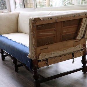 May include: A vintage, white linen sofa with exposed wooden frame and dark brown legs. The sofa is in need of reupholstery, with the fabric torn and the stuffing visible.