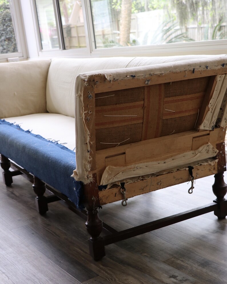 May include: A vintage, white linen sofa with exposed wooden frame and dark brown legs. The sofa is in need of reupholstery, with the fabric torn and the stuffing visible.