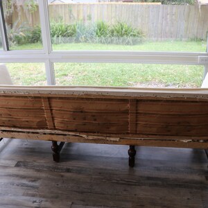May include: The back of a vintage sofa with wooden frame and legs. The fabric is white and the sofa is in need of reupholstery.
