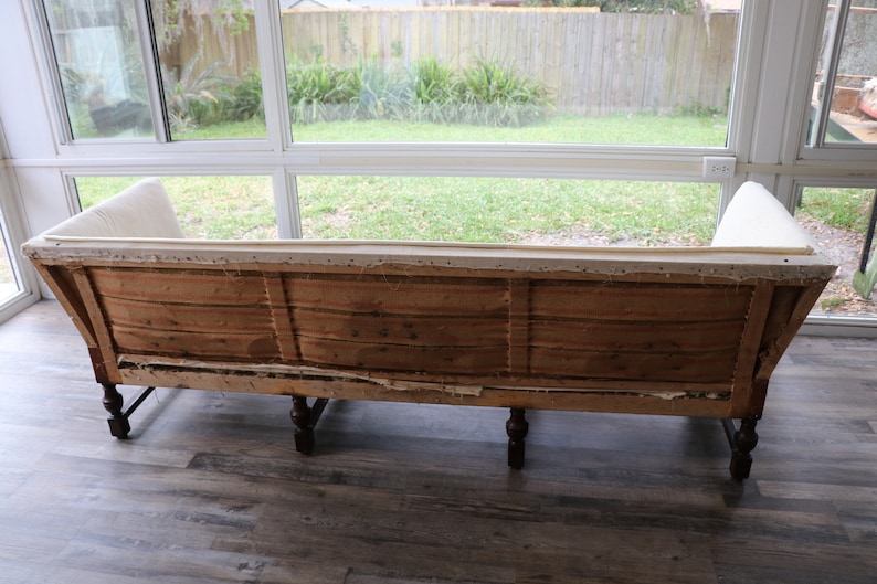 May include: The back of a vintage sofa with wooden frame and legs. The fabric is white and the sofa is in need of reupholstery.