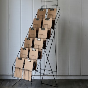 May include: A black metal display rack with multiple clipboards. The clipboards are arranged in a tiered fashion, with each level holding several clipboards. The clipboards have a natural, light brown color, and the metal clips are silver.