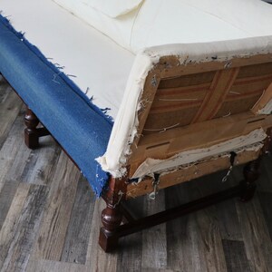 May include: A vintage sofa with a blue denim fabric covering the seat and back cushions. The sofa has a wooden frame and legs, and the back cushion is partially removed, revealing the inner structure and fabric.