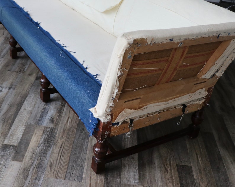 May include: A vintage sofa with a blue denim fabric covering the seat and back cushions. The sofa has a wooden frame and legs, and the back cushion is partially removed, revealing the inner structure and fabric.