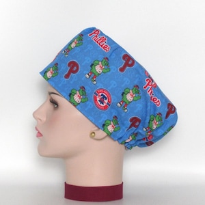 May include: Blue scrub hat with a Phillies baseball team print featuring the team mascot, the Phillie Phanatic. The hat has a red, white, and blue logo with the words "Philadelphia Phillies" on it.