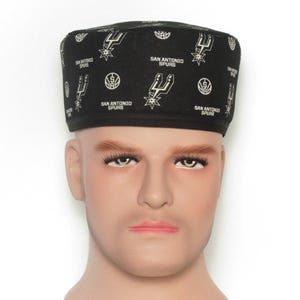 Puede incluir: Gorra negra con el logotipo y el texto de los San Antonio Spurs. La gorra presenta un patr&oacute;n repetido del logotipo del equipo y las palabras "San Antonio Spurs" en blanco. La gorra se muestra en una cabeza de maniqu&iacute;.