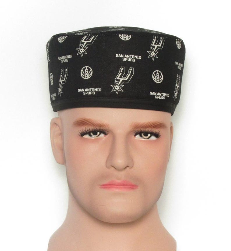 Puede incluir: Gorra negra con el logotipo y el texto de los San Antonio Spurs. La gorra presenta un patr&oacute;n repetido del logotipo del equipo y las palabras "San Antonio Spurs" en blanco. La gorra se muestra en una cabeza de maniqu&iacute;.