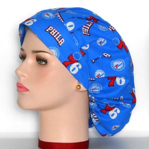 May include: A blue scrub cap featuring the Philadelphia 76ers logo and team name. The cap has a repeating pattern of the team's logo, the number 76, and the word "PHILA" in red and white. The cap is made of a soft, lightweight fabric.