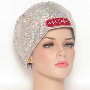 May include: A silver, iridescent surgical scrub cap with a red patch featuring a white heartbeat line and heart symbol. The cap is on a mannequin head, along with a matching choker. The cap has a textured, scale-like pattern.