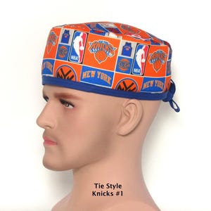 May include: A scrub cap featuring an orange and blue grid pattern with New York Knicks basketball team logos, NBA emblems, and the text New York. The cap has a solid blue fabric band along the bottom edge and includes tie-back strings at the rear.