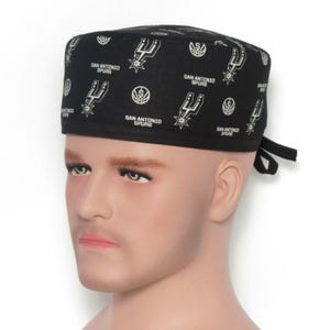 Puede incluir: Gorro quir&uacute;rgico negro con un patr&oacute;n repetido del logotipo y el texto de los San Antonio Spurs en blanco. El gorro presenta el logotipo y el nombre del equipo. Se ata en la parte posterior con lazos negros.