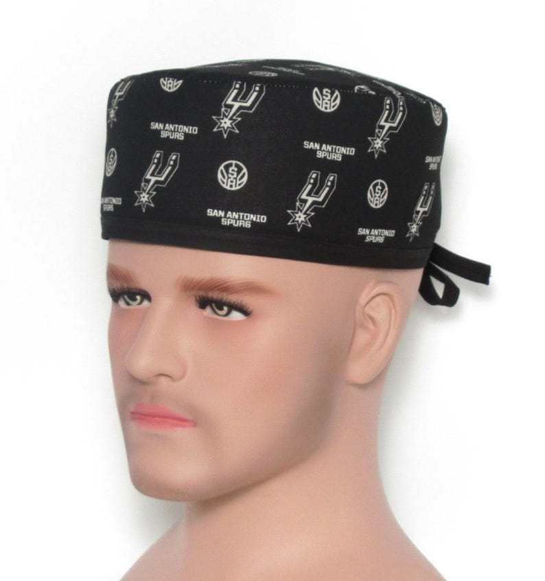 Puede incluir: Gorro quir&uacute;rgico negro con un patr&oacute;n repetido del logotipo y el texto de los San Antonio Spurs en blanco. El gorro presenta el logotipo y el nombre del equipo. Se ata en la parte posterior con lazos negros.