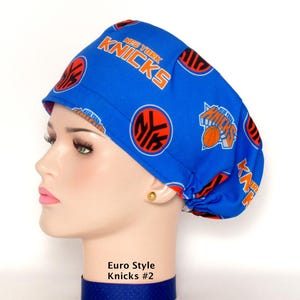 May include: A blue scrub cap featuring a repeating pattern of the New York Knicks basketball team logo and text. The fabric displays the orange and blue team colors with circular emblems and the word Knicks printed in bold orange lettering.