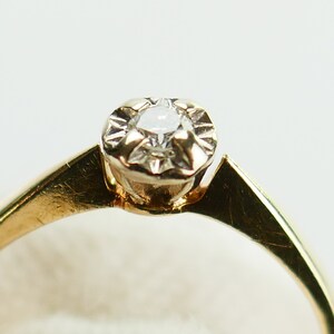 K&ouml;nnte beinhalten: Ein goldener Ring mit einem einzelnen, klaren Diamanten in einer Krappenfassung.