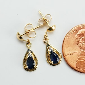 May include: A pair of gold-colored drop earrings with a teardrop-shaped, dark blue gemstone. The earrings have a small, round stud and a decorative border. A penny is next to the earrings for size comparison.